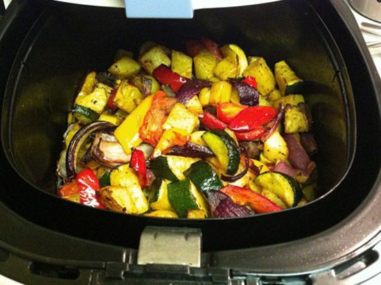 Vista superior do cesto interno de uma fritadeira elétrica (air fryer) repleto de legumes assados e coloridos. É possível identificar pedaços de abobrinha com casca verde, pimentões vermelhos e amarelos, cebola roxa e batatas douradas, todos levemente tostados e temperados, demonstrando o resultado de um cozimento saudável por circulação de ar.
