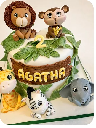 Bolo de 2 anos no tema Safari com leão, macaco com banana, girafa, zebra e elefante todos com expressão baby, letreiro com nome do aniversário na frente do bolo de 1 andar e folhas do Curso Meu Primeiro Zoológico - Modele 25 Animais em Pasta Americana