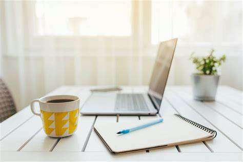 Uma mesa com notebook aberto, e ao lado, um caderno com uma caneta para fazer anotações e uma xícara de café, e um delicado vaso com planta ao fundo, indicando que o cenário está pronto para quem quiser começar a estudar.