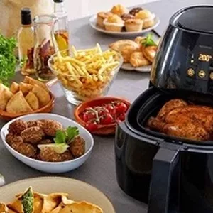 Mesa farta de jantar com diversos petiscos ao lado de uma air fryer preta digital aberta, revelando um frango assado inteiro em seu interior. Sobre a mesa, veem-se tigelas com batatas fritas palito, almôndegas, batatas rústicas, tomates cereja e muffins, ilustrando a capacidade de preparar uma refeição completa com o aparelho.