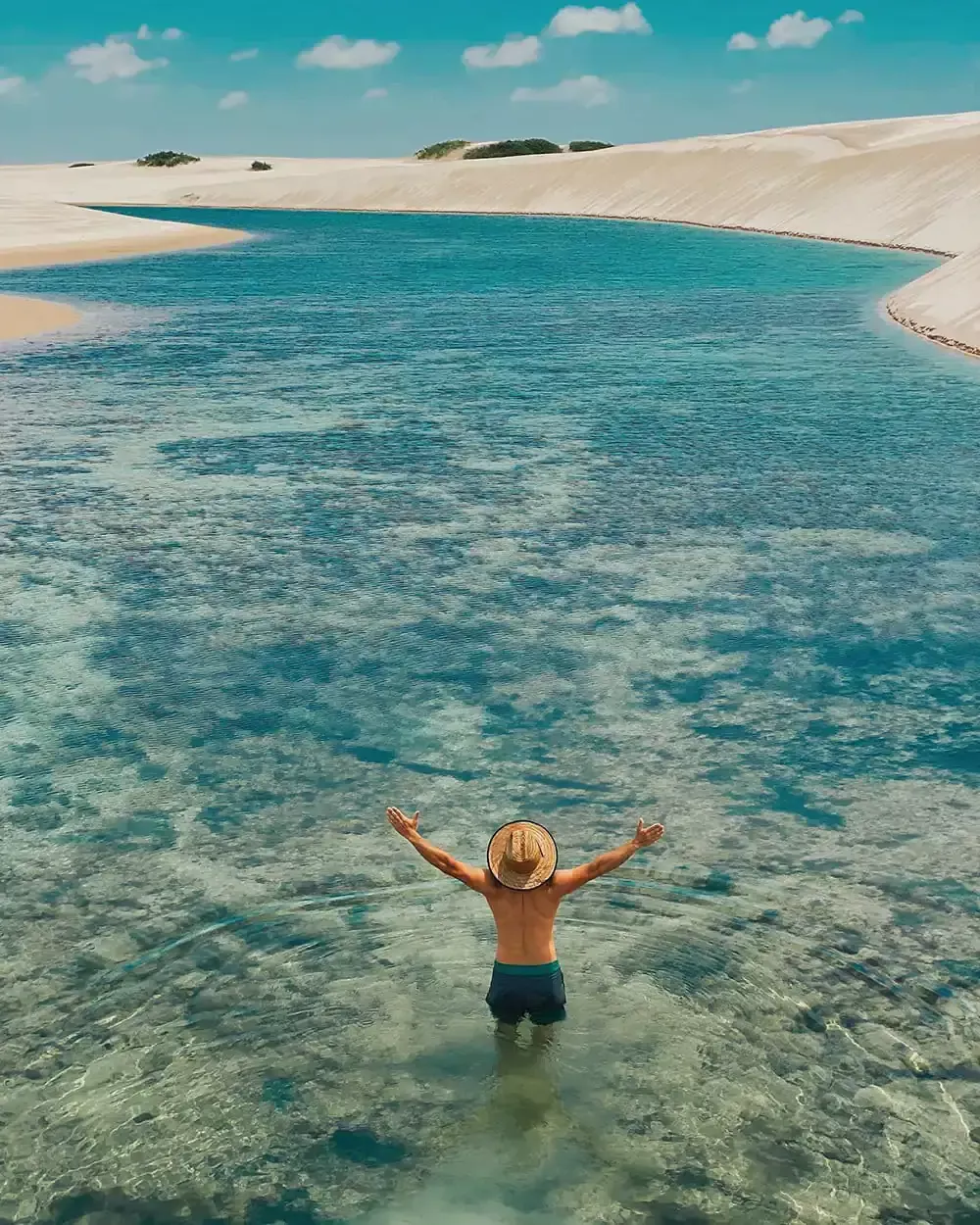 The photo shows a man from behind, standing in the shallow water of a crystalline lagoon, with his arms open in a welcoming gesture. He wears a straw hat and swimming shorts, with most of his back and legs visible. The water is so clear that you can see the sandy bottom and the soft ripples created by his feet. The lagoon extends towards the horizon, with the water varying from a deep blue to a light turquoise, depending on the depth and light. The waters surface reflects the clear blue sky, which is dotted with a few white clouds. In the background, there are white sand dunes that stretch as far as the eye can see, creating a desert landscape contrasting with the lagoons water. The clear blue sky and white clouds create a serene backdrop for the scene. The photo is taken from a high angle, showing the vastness of the landscape and the smallness of the human figure in relation to the natural environment. Natural light illuminates the scene, creating soft reflections on the water and highlighting the texture of the sand and clouds. Additional Elements: Colors: Predominantly blue (water and sky) and white (sand and clouds). Composition: The man is centered in the photo, with the lagoon and sand dunes extending towards the horizon. Atmosphere: Serene, inviting, and contemplative.