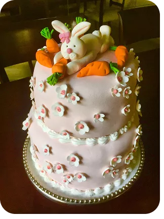 Bolo de 2 andares no tema Páscoa com cores suaves, no topo do bolo uma coelha comendo uma cenoura e cenouras, na base flores por todo o bolo do Curso Meu Primeiro Zoológico - Modele 25 Animais em Pasta Americana