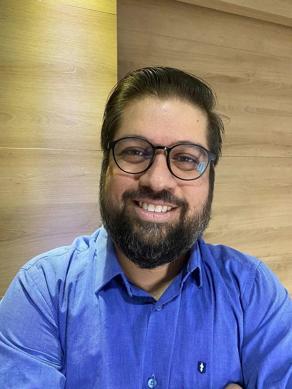Homem adulto sorrindo, com barba e óculos de armação escura, usando camisa social azul, em frente a uma parede de madeira clara.A foto é um retrato em pé (formato vertical), enquadrado do peito para cima. Um homem aparece no centro, olhando para a câmera e sorrindo, com os dentes à mostra. Ele usa óculos redondos/ovais de armação preta e tem barba cheia e bem aparada. O cabelo é escuro, penteado para trás, com risca lateral. Ele veste uma camisa social azul de manga comprida, com botões na frente, e está com os braços cruzados. Ao fundo, há uma parede de madeira em tons claros, com placas horizontais e variações de cor; a iluminação é suave e quente, dando um clima bem aconchegante.