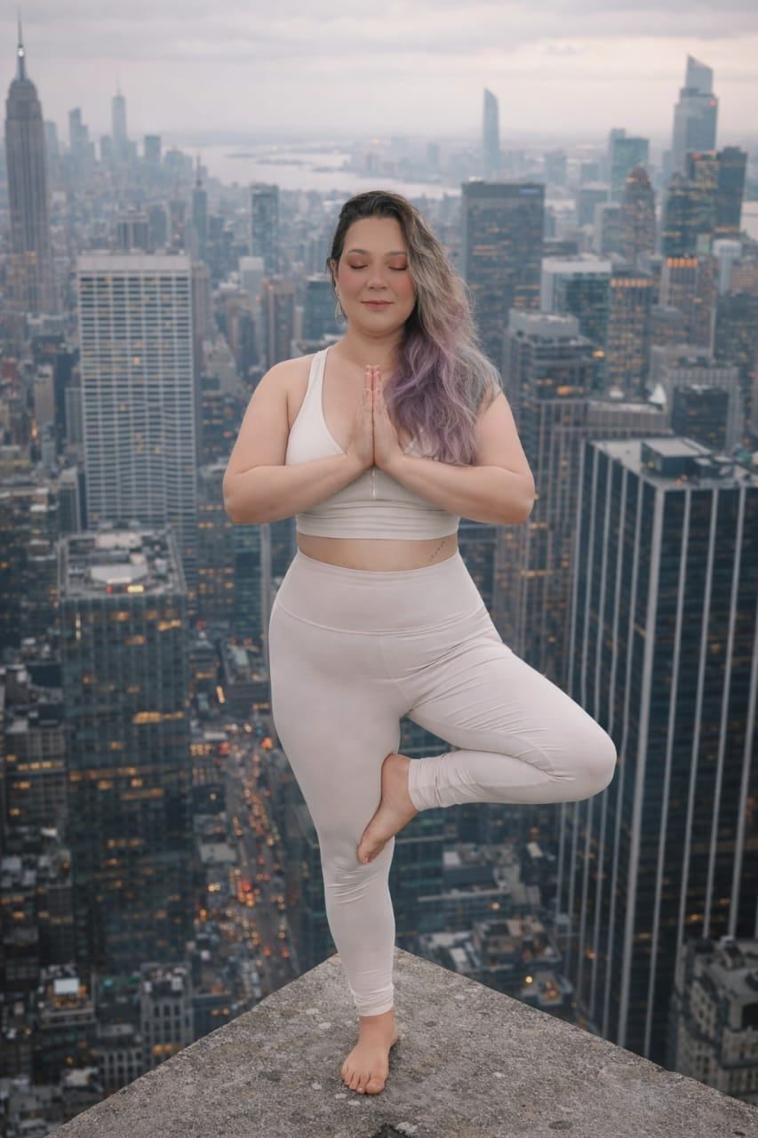 Mulher de cabelos longos com mechas roxas, usando roupa clara, em pé fazendo yoga no topo de um prédio alto.
