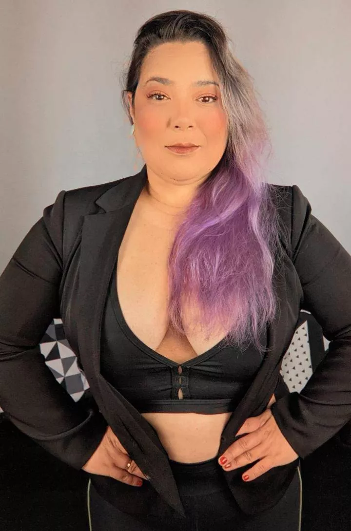 Uma mulher de cabelo roxo, vestindo uma roupa preta, de pé com a mão na cintura com atitude de quem sabe do que está falando.